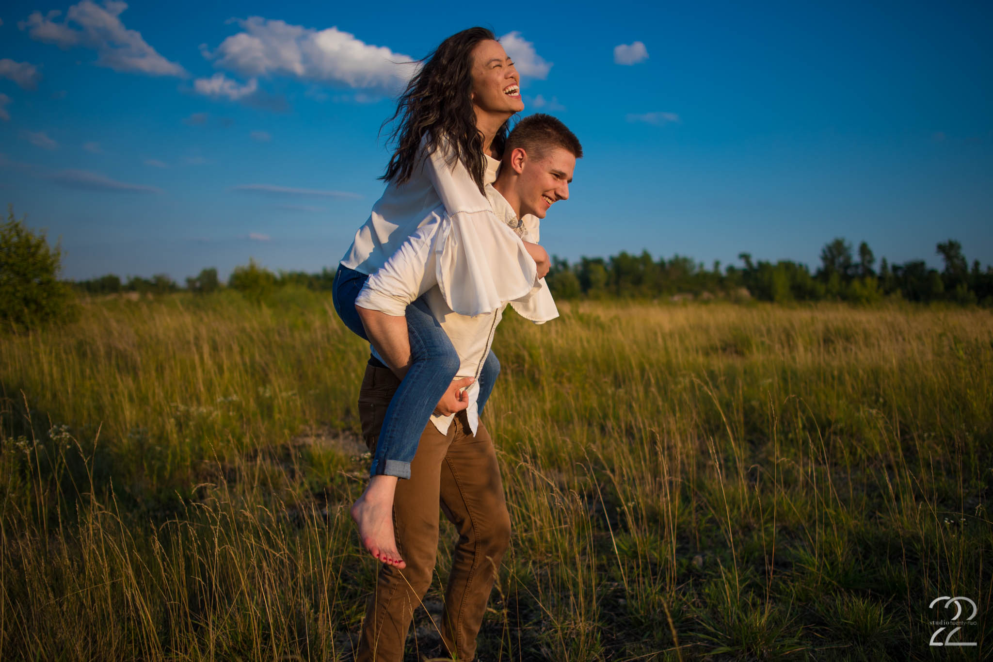 Woman rides piggy back on a man's back, laughing as they run through a field with a blue sky behind them at Oakes Quarry Park by Dayton Wedding Photographer Studio 22 Photography